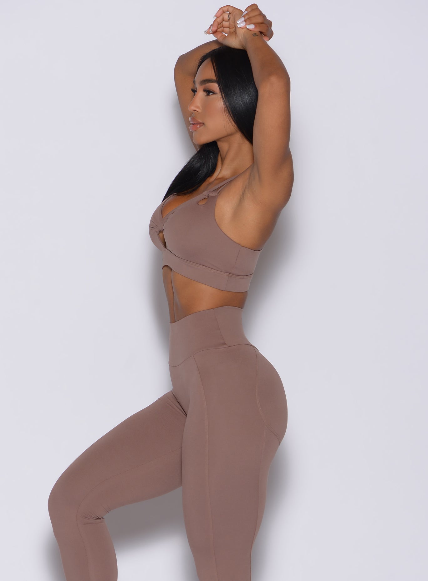 Left side profile view of a model with both her hand over her head wearing our twist sports bra in tan color and a matching leggings