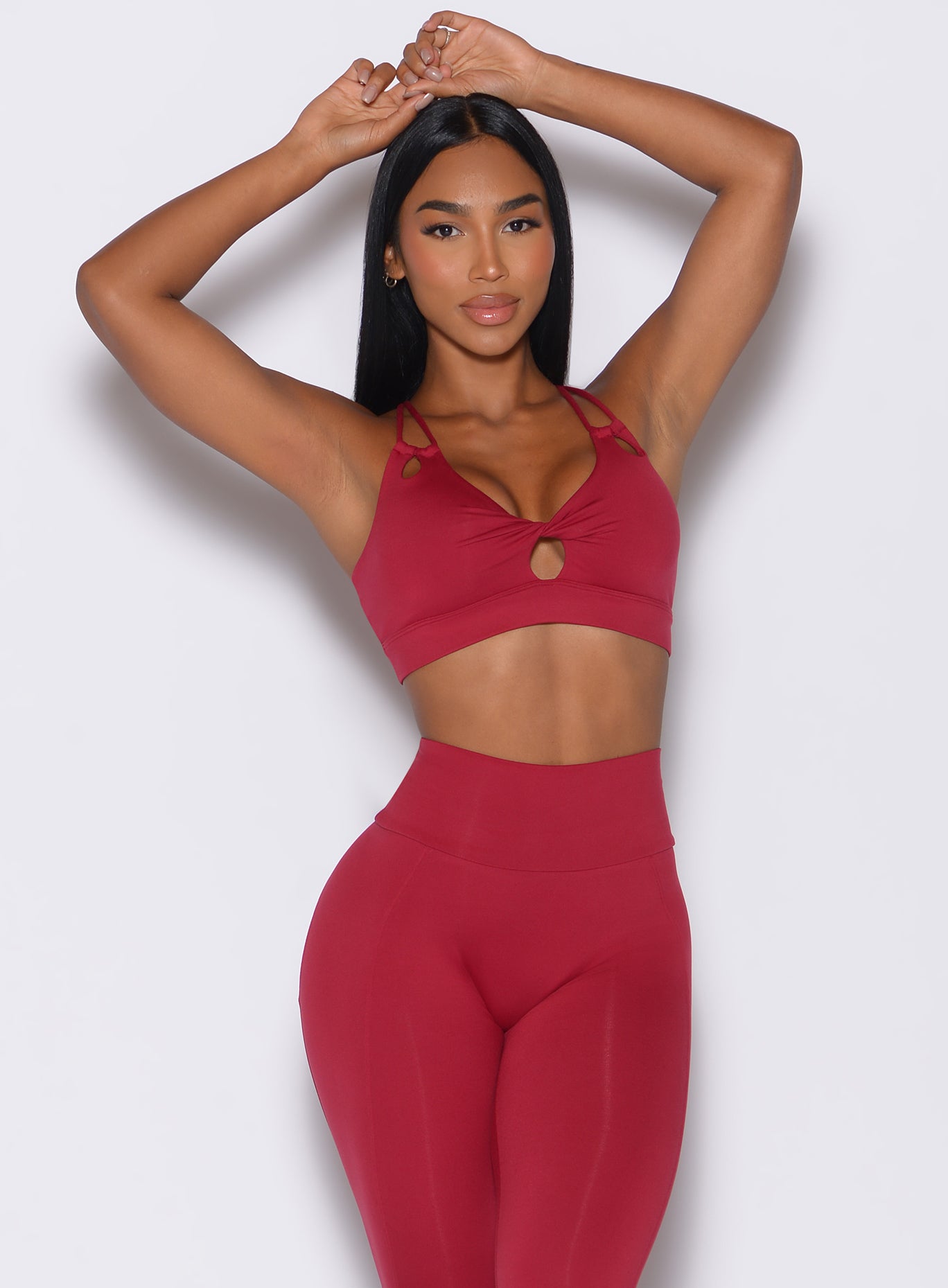 Front profile view of a model with her hands over her head wearing our twist sports bra in maroon color along with a matching leggings