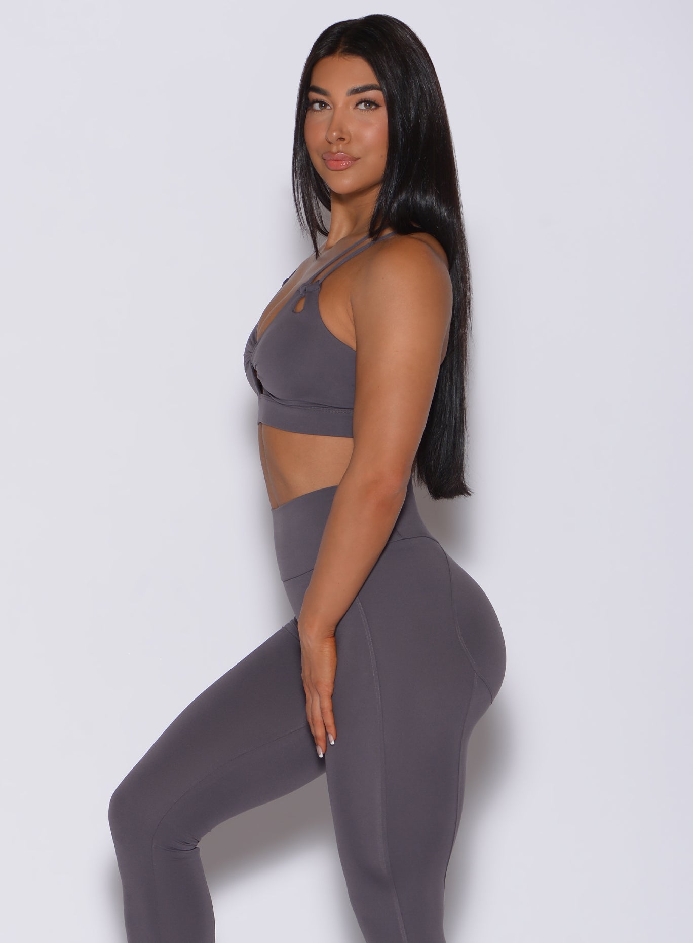 Left side profile view of a model facing to her left wearing our twist sports bra in gray smoke color and a matching leggings
