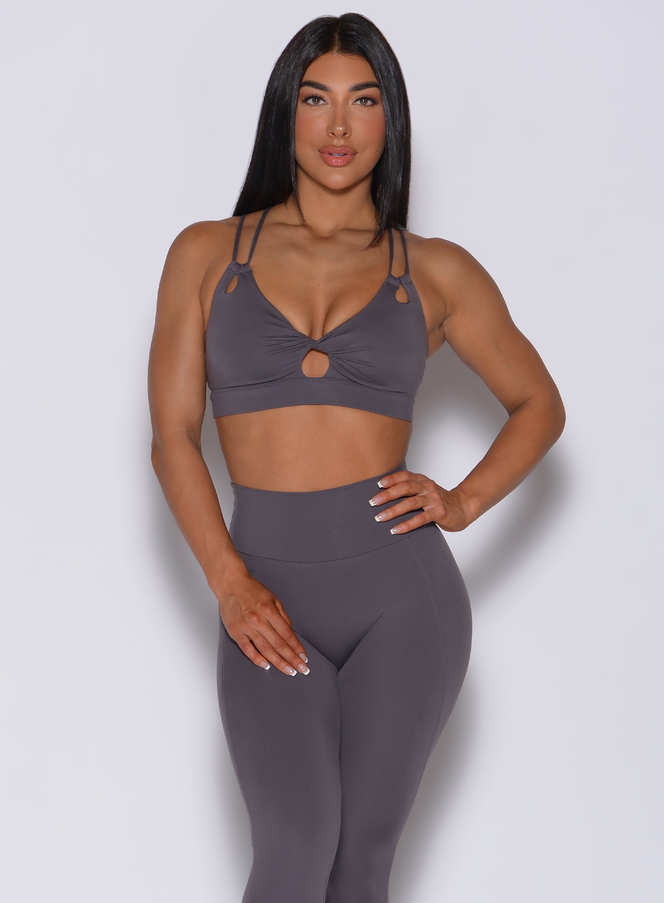 Front profile view of a model with her left hand on waist wearing our twist sports bra in gray smoke color and a matching leggings