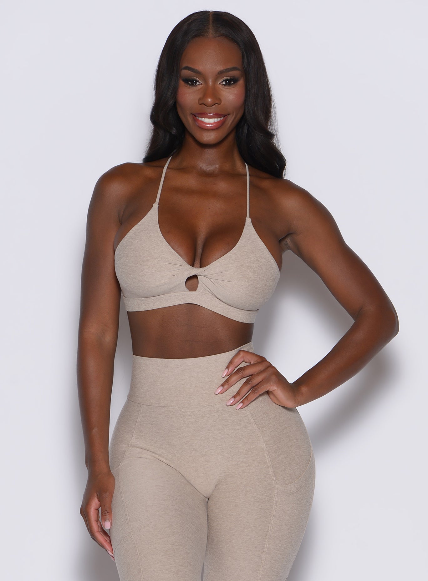 front profile view of a model with her left hand on waist wearing our tiny twist bra in taupe color along with the matching curves leggings
