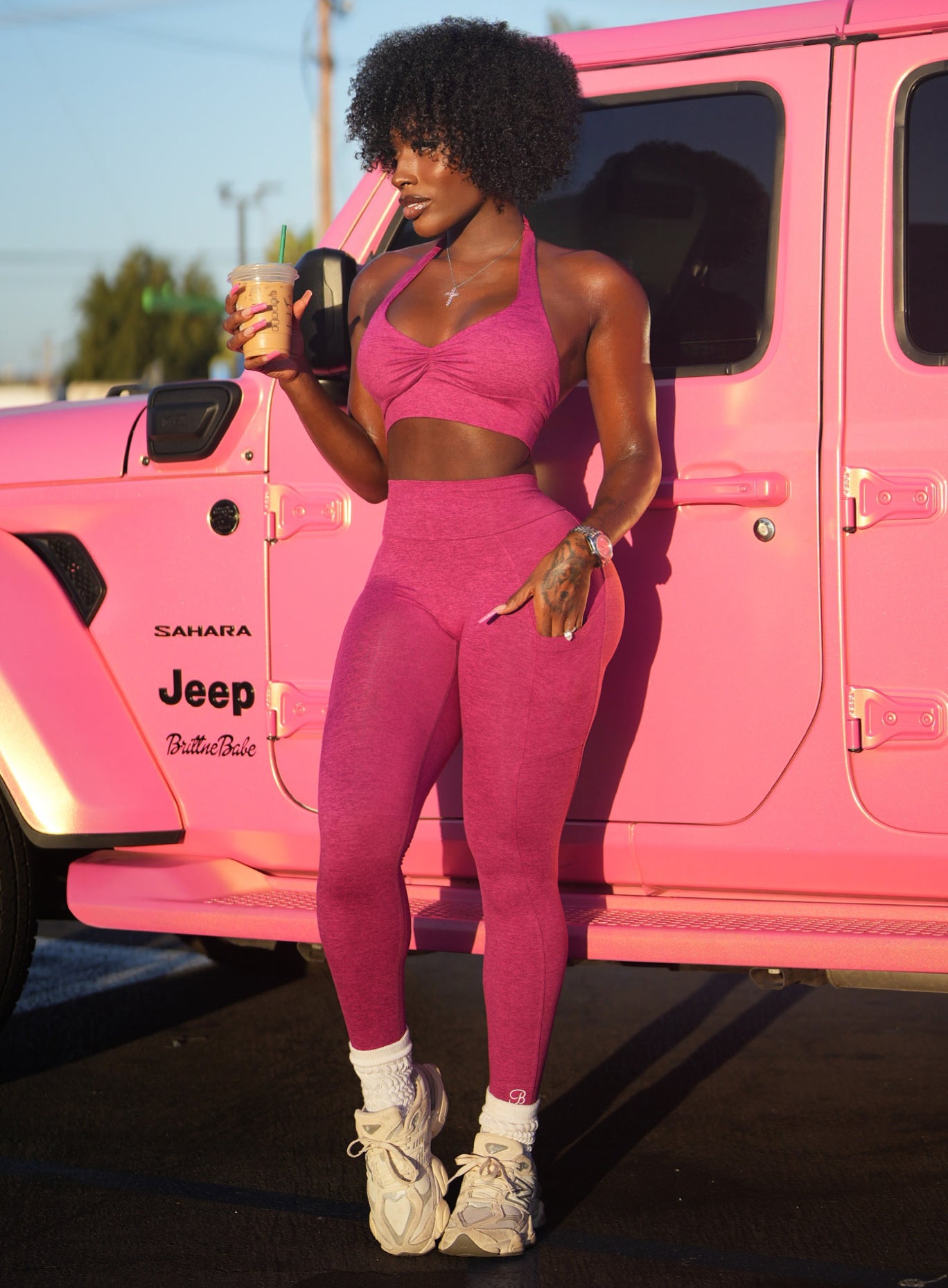 front side profile view of model wearing the fuchsia v back leggings and backless bra standing outside by a pink jeep holding a coffee