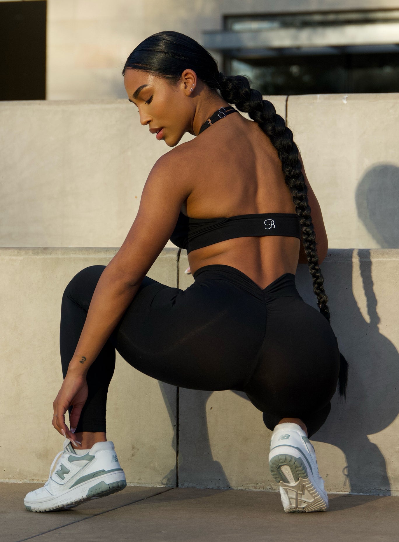 Back view of a model sitting in squat position adjusting her shoes wearing our black backless bra along with the  matching leggings 