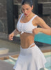 Video of woman wearing a white bikini set with a knotted tank-style bikini top and strappy bottoms, paired with a matching mini skirt, posing poolside.