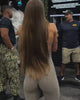 A woman, wearing a tan jumpsuit that clearly show her back muscles, actively doing pull ups in an indoor gym space. 