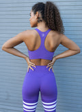 Back view of the model with her hands on waist wearing  our perform thigh highs in royal purple color with three white stripes on thighs and a matching bra