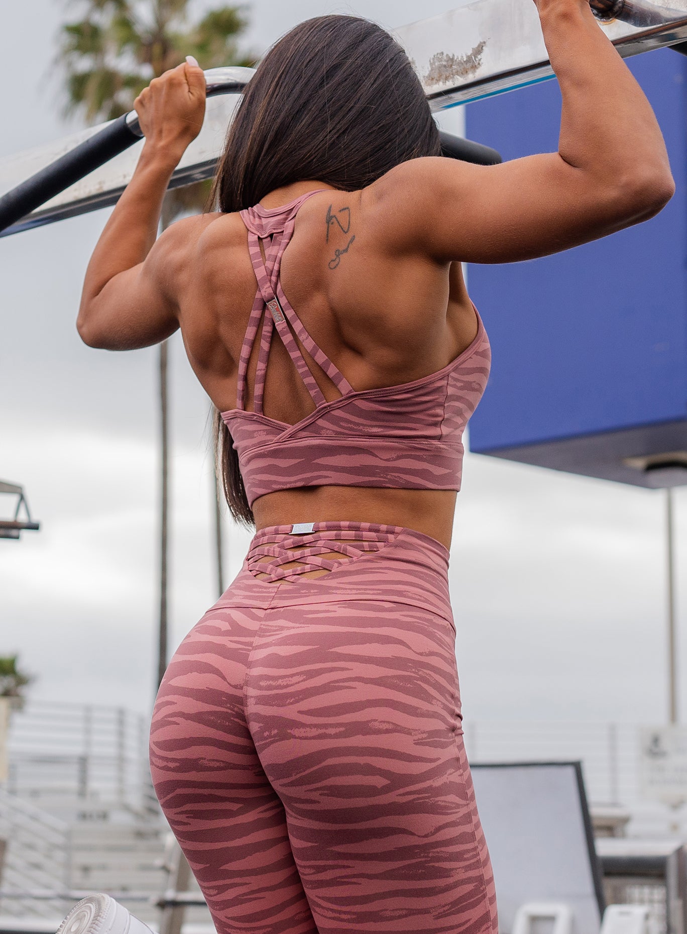 Back profile view of a model doing pull ups and showing off her muscles wearing our rival sports bra in nude blush color and a matching leggings 